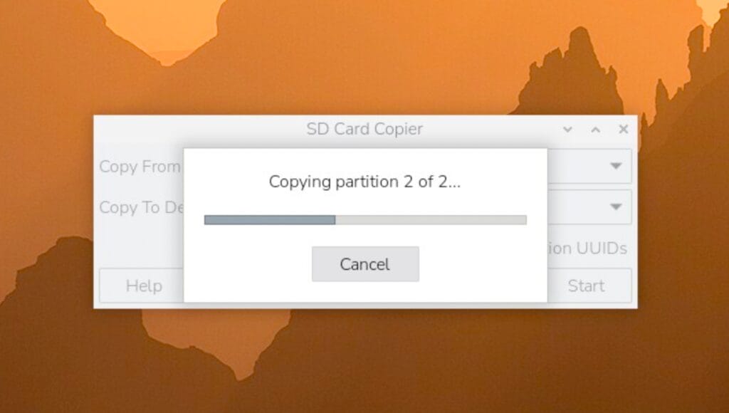 Im Vordergrund ist ein Dialogfenster der Software SD Card Copier des Betriebssystems Raspberry Pi OS zu sehen. Das Fenster zeigt einen Fortschrittsbalken mit der Beschriftung 'Copying partition 2 of 2...'. Darunter befindet sich eine Schaltfläche mit der Aufschrift 'Cancel'. Im Hintergrund ist unscharf ein orangefarbener Desktop-Hintergrund mit Bergsilhouetten zu erkennen.