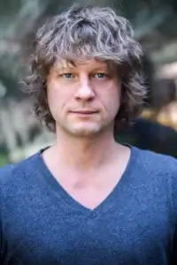 Eine Person mit mittellangem, lockigem, braunem Haar und blauen Augen blickt direkt in die Kamera. Sie trägt ein dunkelblaues T-Shirt mit V-Ausschnitt. Der Hintergrund ist unscharf und zeigt grüne und graue Farbtöne, was auf eine Außenaufnahme schließen lässt. Die Gesichtszüge sind neutral und entspannt.